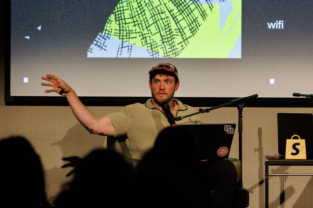 Dom mid-explanation on stage, laptop on lap, hand in the air
